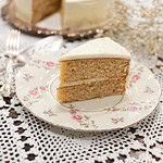 Overhead view of a frosted sourdough vanilla layer cake on a silver platter with a slice removed, served on a vintage floral plate beside baby’s breath flowers.
