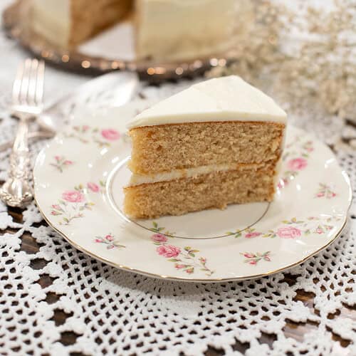 Overhead view of a frosted sourdough vanilla layer cake on a silver platter with a slice removed, served on a vintage floral plate beside baby’s breath flowers.