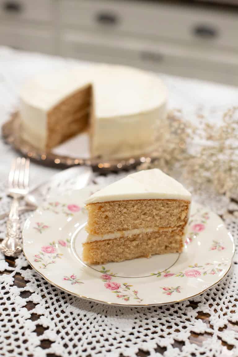 Overhead view of a frosted sourdough vanilla layer cake on a silver platter with a slice removed, served on a vintage floral plate beside baby’s breath flowers.