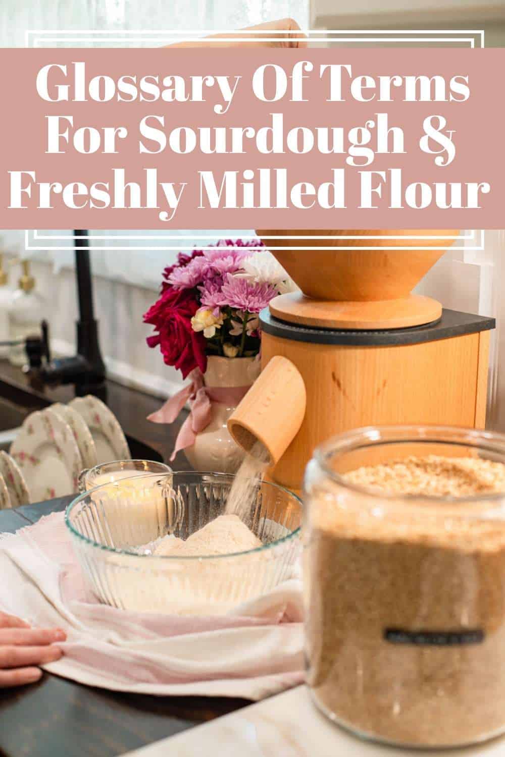 Wooden grain mill pouring freshly milled flour into a glass bowl with a jar of wheat berries and flowers in the background.