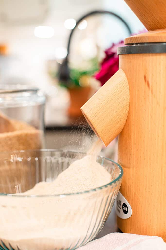 Close-up of a wooden grain mill grinding flour into a glass bowl on a kitchen counter.