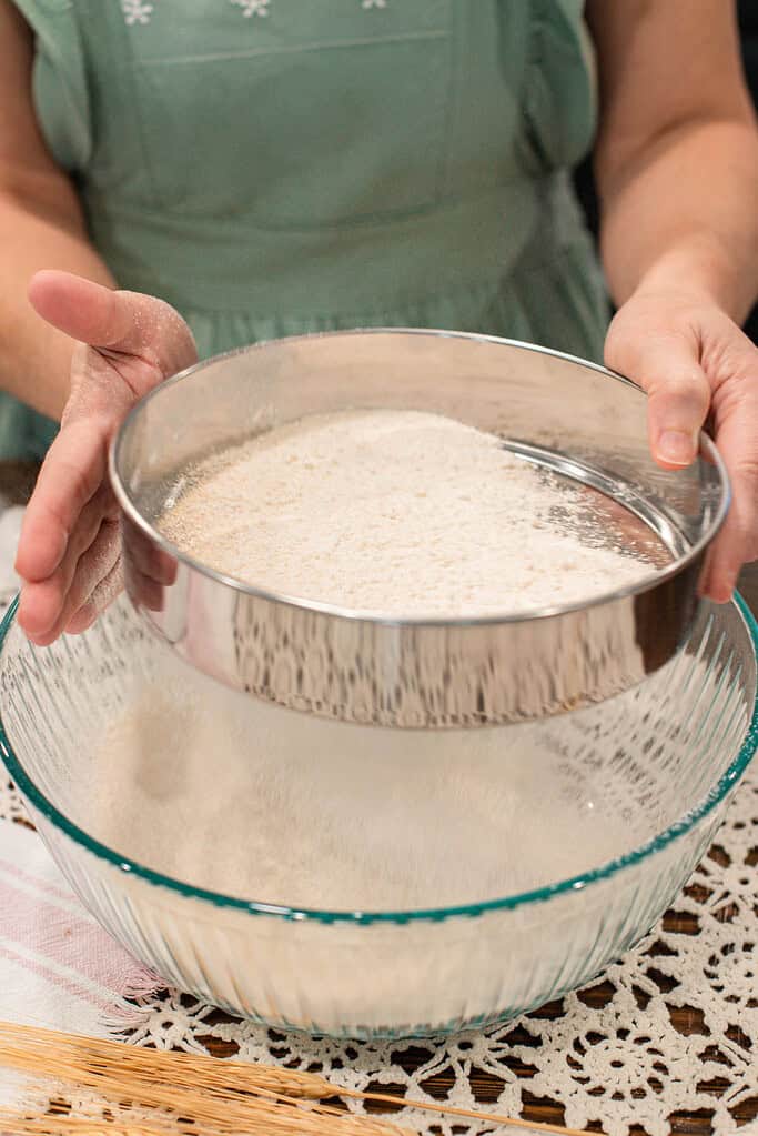 Hands sifting freshly milled flour through a metal sieve into a glass bowl on a lace-covered table.