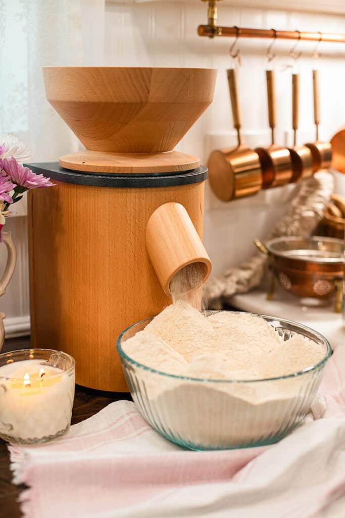A wooden flour mill dispensing freshly ground flour into a glass bowl in a cozy kitchen with copper utensils.