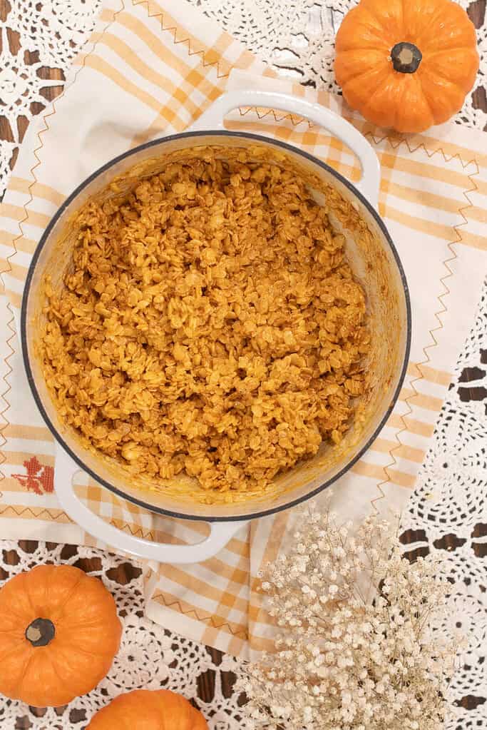 Pumpkin spice rice krispie mixture stirred together in a white Dutch oven, sitting on a yellow plaid towel with mini pumpkins nearby.