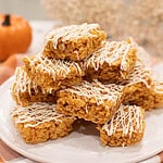 A stack of pumpkin spice rice krispie treats drizzled with white chocolate, arranged on a white plate with a soft fall background.