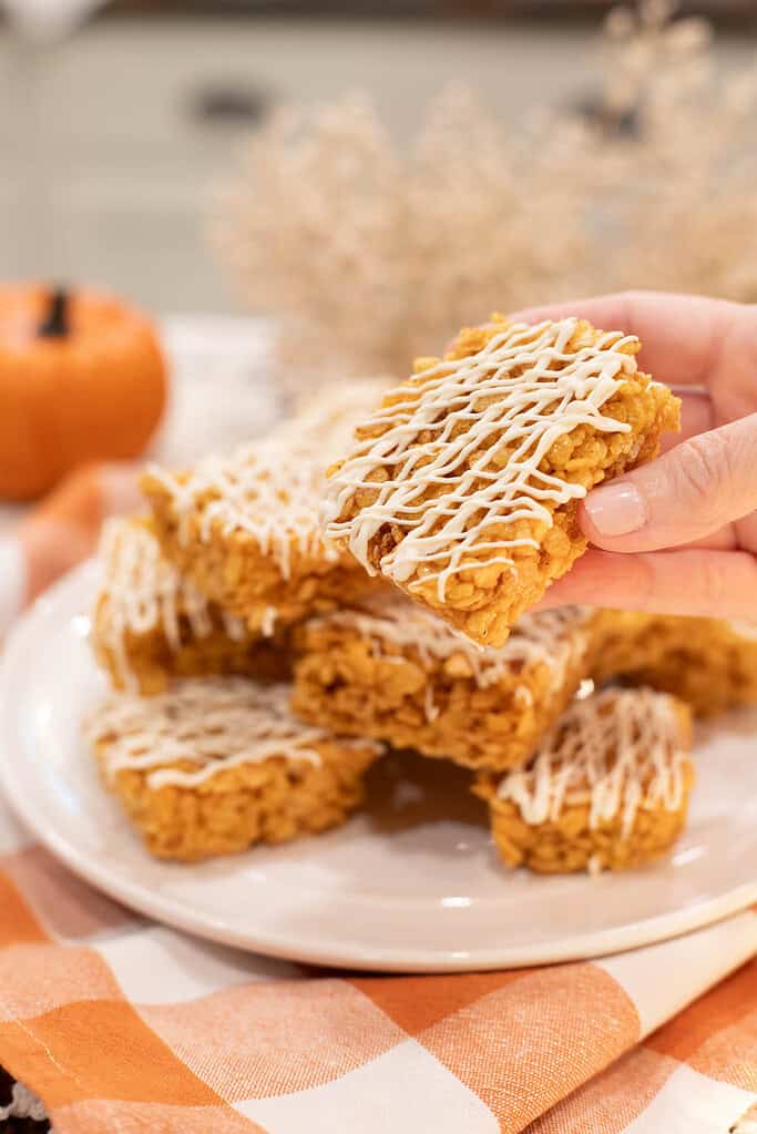 A hand holding a pumpkin spice rice krispie treat drizzled with white chocolate, with more bars stacked on a plate in the background.