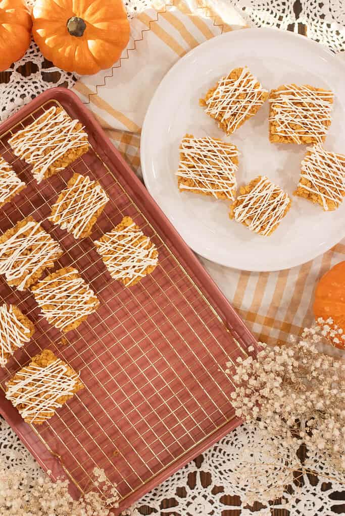 Overhead view of pumpkin spice rice krispie treats cooling on a rack beside a white plate with five bars, styled with pumpkins, lace, and dried flowers for fall.