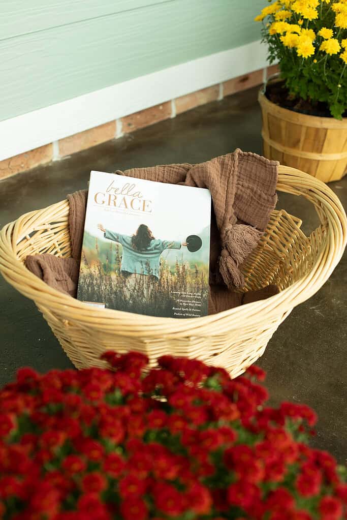 Cozy fall porch basket with a brown throw blanket and Bella Grace magazine beside red and yellow mums.