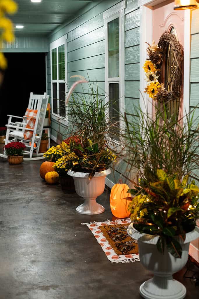 A cozy cottage-style front porch decorated for fall, featuring white rocking chairs with orange plaid pillows, potted mums, and pumpkins arranged along the walkway. A peach-colored front door with a sunflower wreath and a softly glowing pumpkin lantern add warmth to the mint-green siding, creating a welcoming autumn glow under the porch lights.