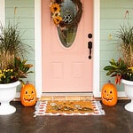 Peach-colored front door decorated for fall with a sunflower wreath, jack-o-lantern pumpkins, and autumn planters.