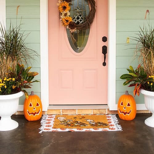 Peach-colored front door decorated for fall with a sunflower wreath, jack-o-lantern pumpkins, and autumn planters.