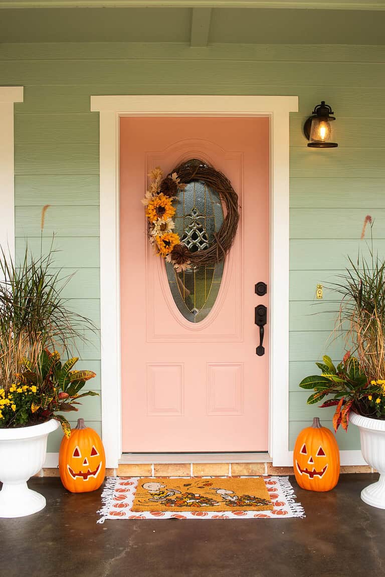 Peach-colored front door decorated for fall with a sunflower wreath, jack-o-lantern pumpkins, and autumn planters.