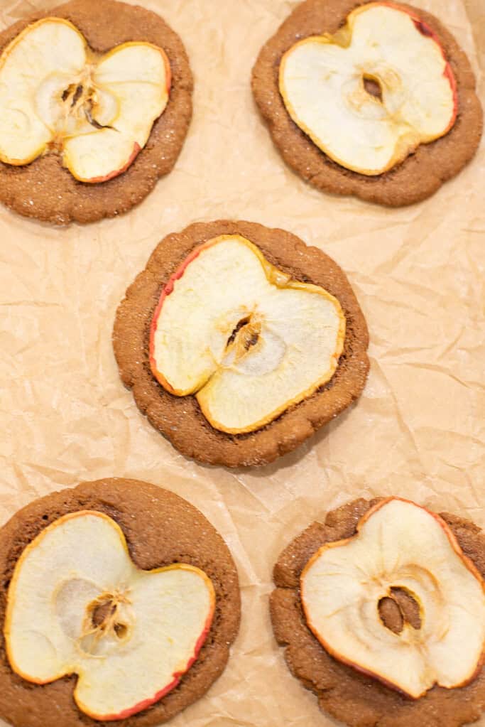Freshly baked nutmeg ginger apple snap cookies topped with dried apple slices on brown parchment paper.