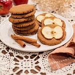 Plate of Mrs. Bean’s nutmeg ginger apple snaps stacked beside cinnamon sticks and fresh apples on a lace tablecloth.