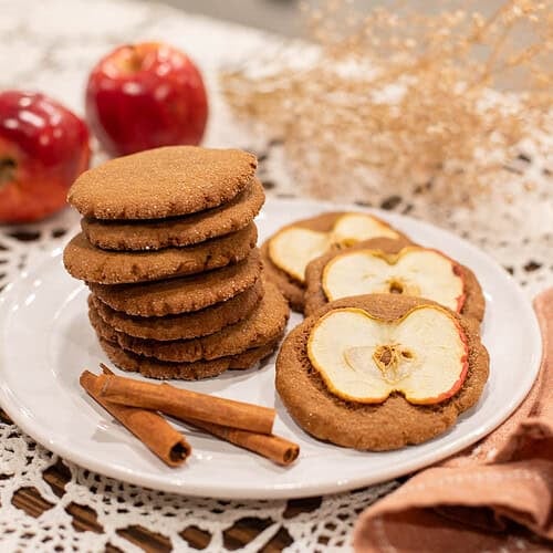 Plate of Mrs. Bean’s nutmeg ginger apple snaps stacked beside cinnamon sticks and fresh apples on a lace tablecloth.