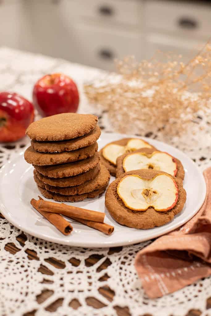 Plate of Mrs. Bean’s nutmeg ginger apple snaps stacked beside cinnamon sticks and fresh apples on a lace tablecloth.