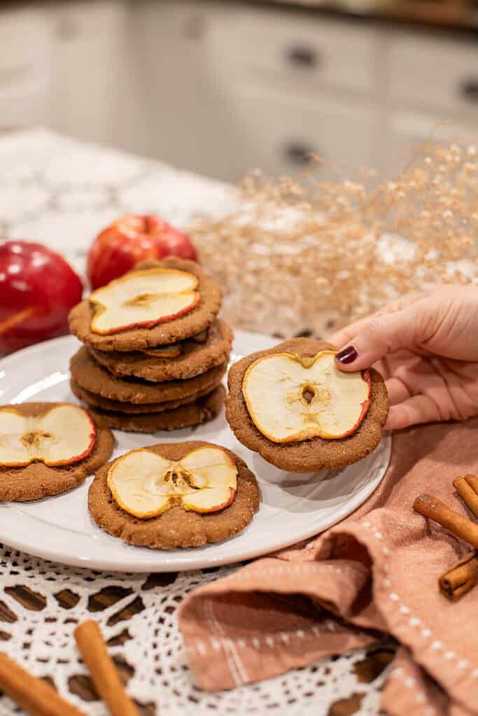 Hand holding a nutmeg ginger apple snap cookie topped with a dried apple slice, with more cookies and red apples in the background.