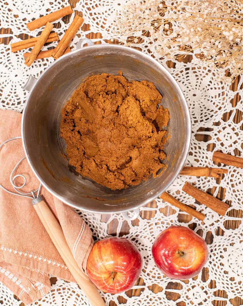 Mixing bowl of spiced ginger snap dough surrounded by apples, cinnamon sticks, and a linen napkin on a lace-covered wooden table.