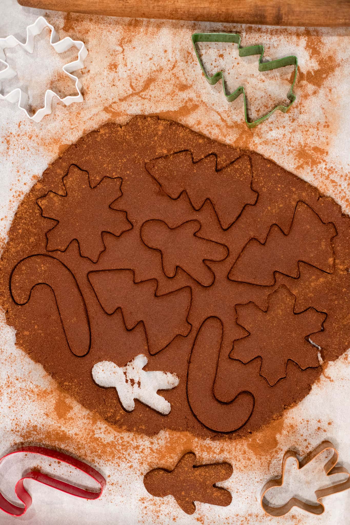 Rolled-out cinnamon dough with Christmas-shaped cookie cutters—gingerbread men, trees, snowflakes, and candy canes—ready to cut on parchment paper dusted with cinnamon.
