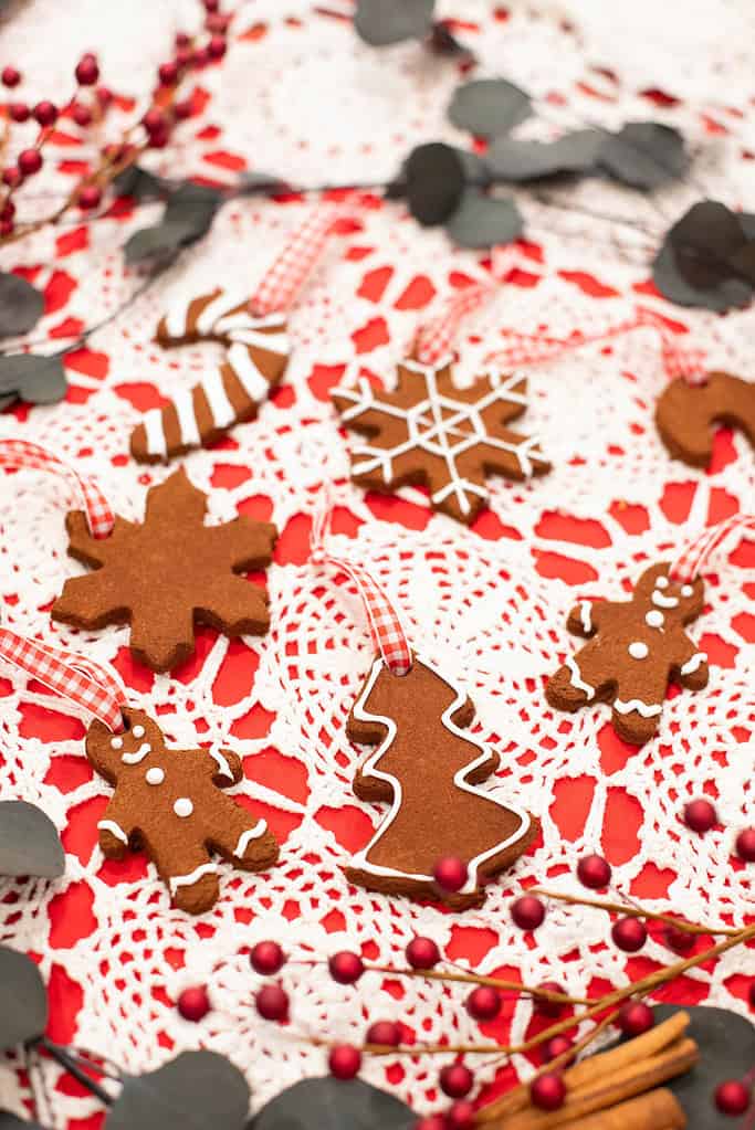 Finished cinnamon applesauce ornaments decorated with white icing and red gingham ribbons on a red and white crochet tablecloth surrounded by eucalyptus and red berries.