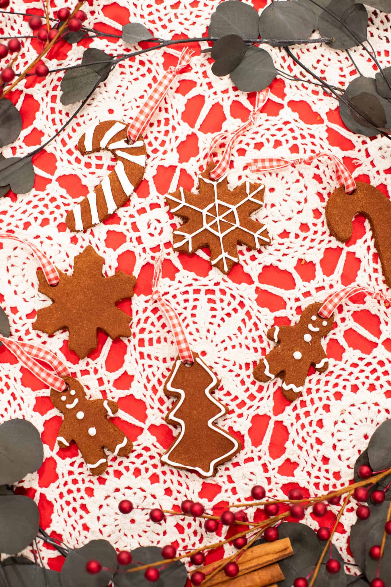 Flat lay of handmade cinnamon applesauce Christmas ornaments in shapes of snowflakes, trees, gingerbread men, and candy canes arranged with red ribbons and holiday greenery.