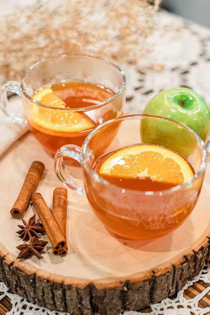 Two glass mugs of homemade apple cider with orange slices, cinnamon sticks, and star anise on a rustic wooden tray beside a green apple.