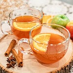 Warm apple cider served in clear mugs with orange slices and cinnamon sticks, displayed on a wooden tray with apples and spices.