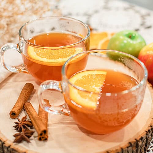 Warm apple cider served in clear mugs with orange slices and cinnamon sticks, displayed on a wooden tray with apples and spices.