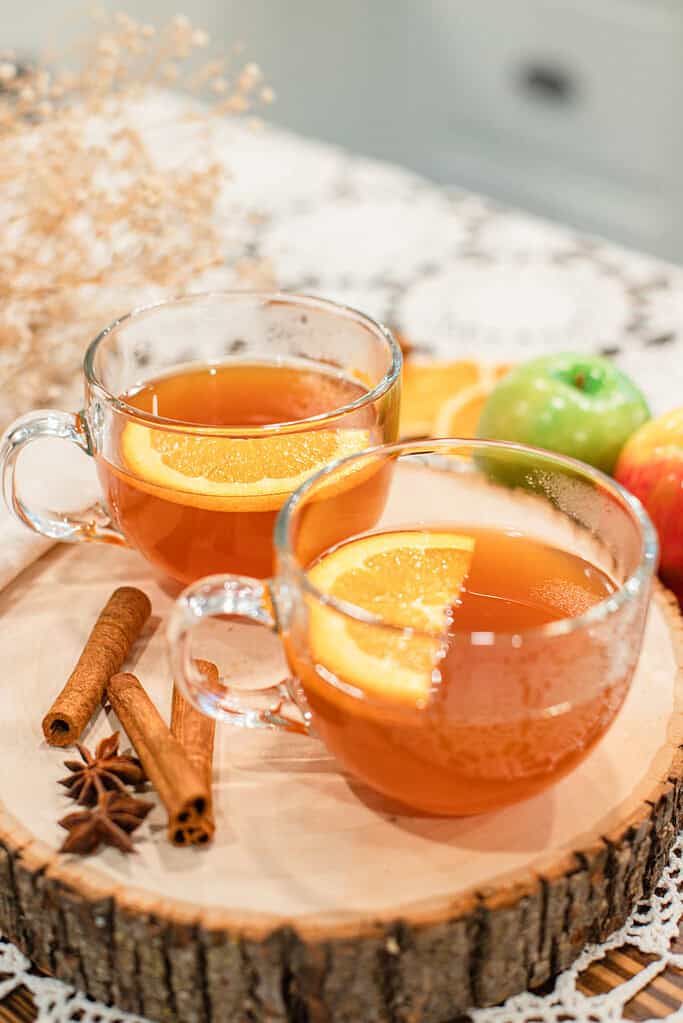 Warm apple cider served in clear mugs with orange slices and cinnamon sticks, displayed on a wooden tray with apples and spices.