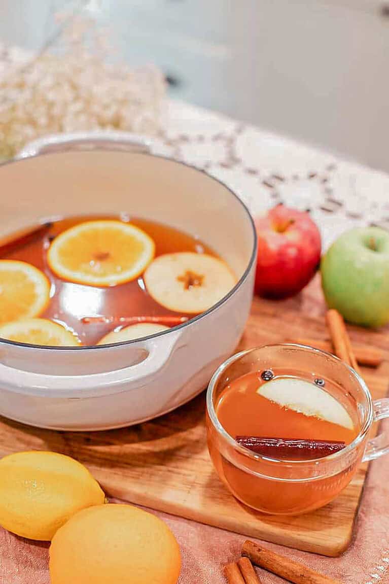 Dutch oven filled with homemade apple cider garnished with apple and orange slices, cinnamon sticks, and star anise, next to a glass mug of cider.