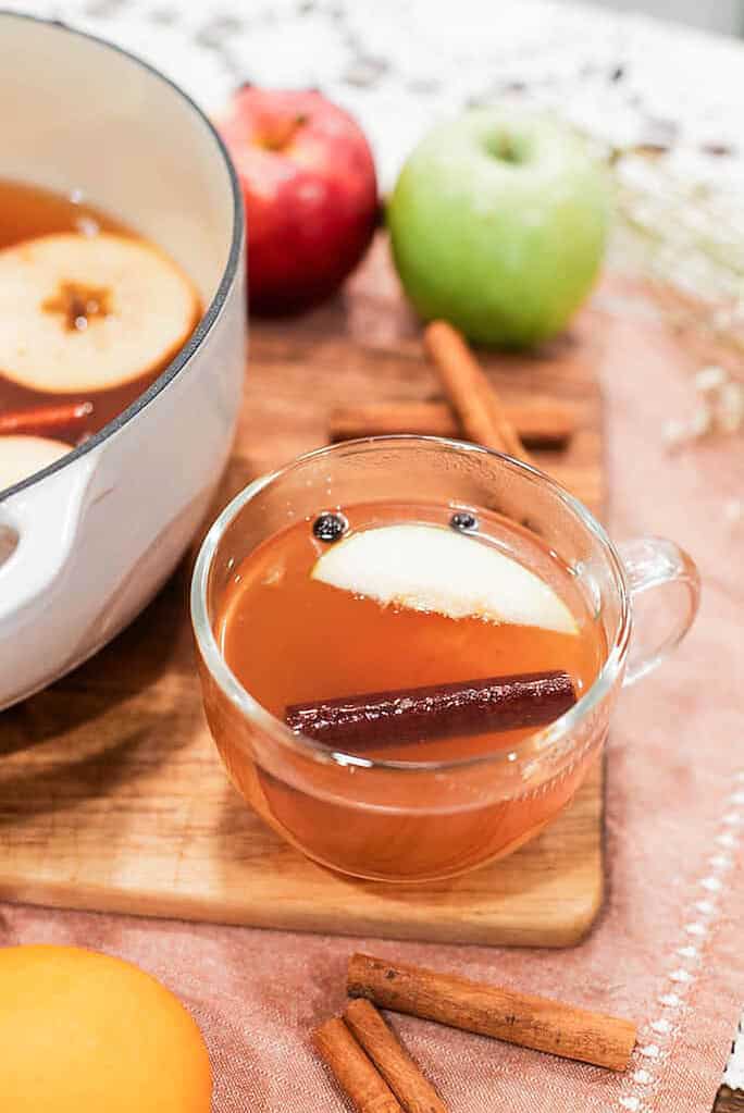 Glass mug of hot apple cider with apple slice and cinnamon stick beside a Dutch oven, surrounded by fresh apples and fall spices.
