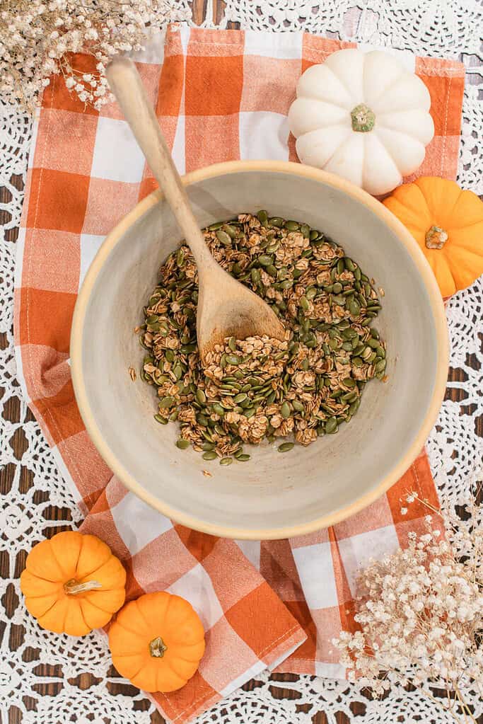 A mixing bowl filled with oats and pumpkin seeds being stirred with a wooden spoon, set on an orange and white checkered cloth with small pumpkins and dried flowers around it.