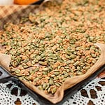 A baking sheet lined with parchment paper holds a spread of golden-brown pumpkin seed brittle, with mini pumpkins blurred in the background.