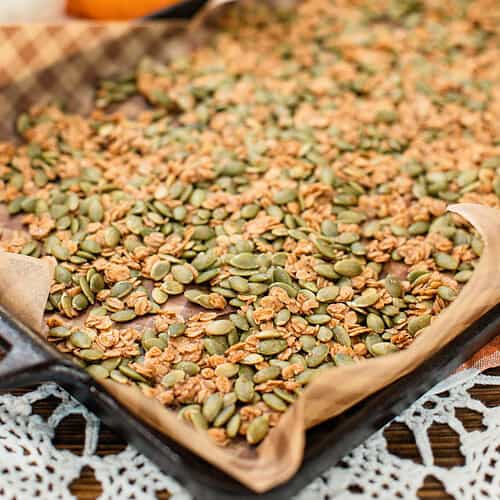 A baking sheet lined with parchment paper holds a spread of golden-brown pumpkin seed brittle, with mini pumpkins blurred in the background.