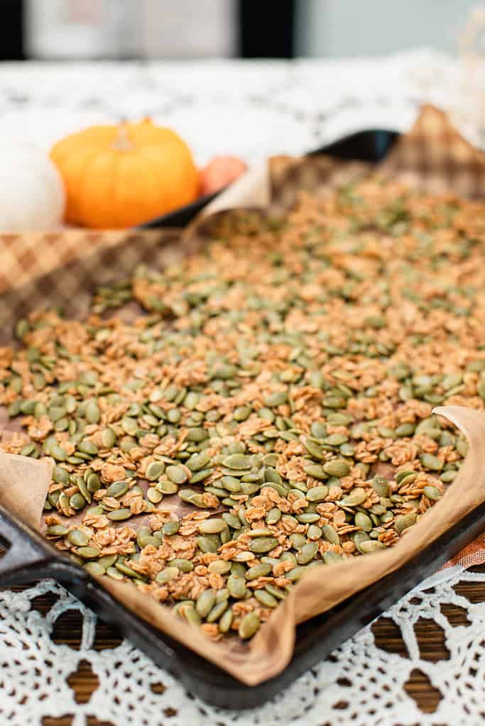 A baking sheet lined with parchment paper holds a spread of golden-brown pumpkin seed brittle, with mini pumpkins blurred in the background.