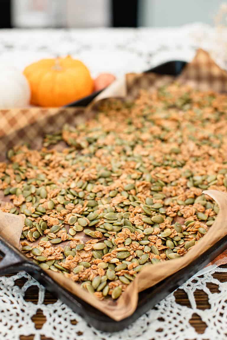 A baking sheet lined with parchment paper holds a spread of golden-brown pumpkin seed brittle, with mini pumpkins blurred in the background.