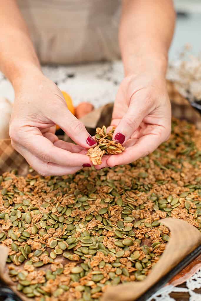 Close-up of hands breaking apart a cluster of pumpkin seed brittle over the baking sheet, showing the crunchy texture.