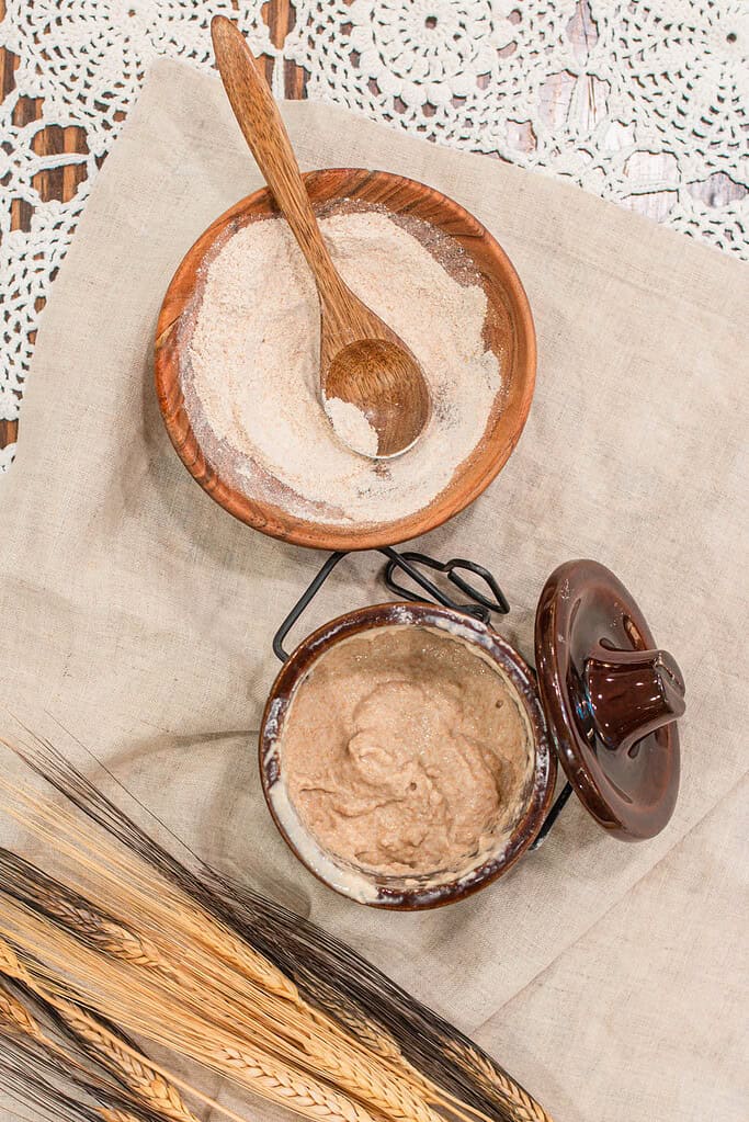 Freshly milled flour and active sourdough starter in rustic brown bowls on a linen cloth with wheat stalks and lace backdrop.
