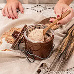 Woman stirring bubbly sourdough starter in a brown crock with a wooden spoon, surrounded by wheat berries and stalks.