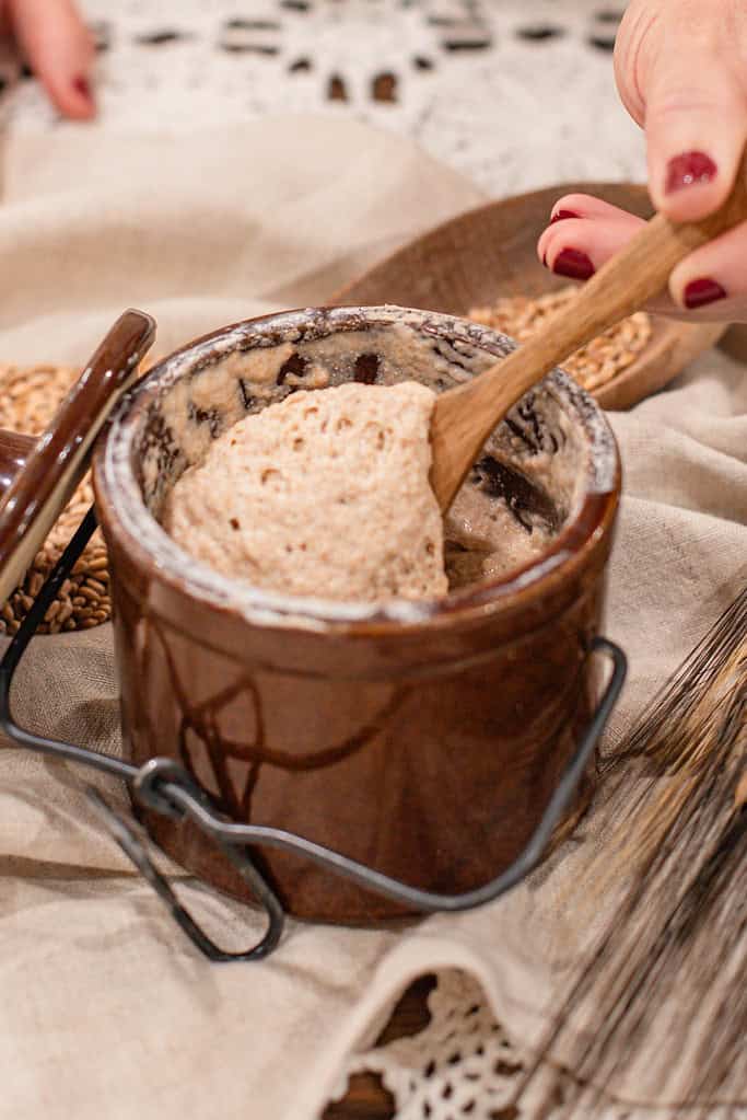 Close-up of airy, active sourdough starter in a brown crock being stirred with a wooden spoon on a cozy farmhouse linen setting.