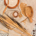 A rustic wooden bowl filled with freshly milled flour, surrounded by golden wheat kernels, stalks of wheat, a wooden scoop, and small bowls of salt and baking powder on a linen cloth.