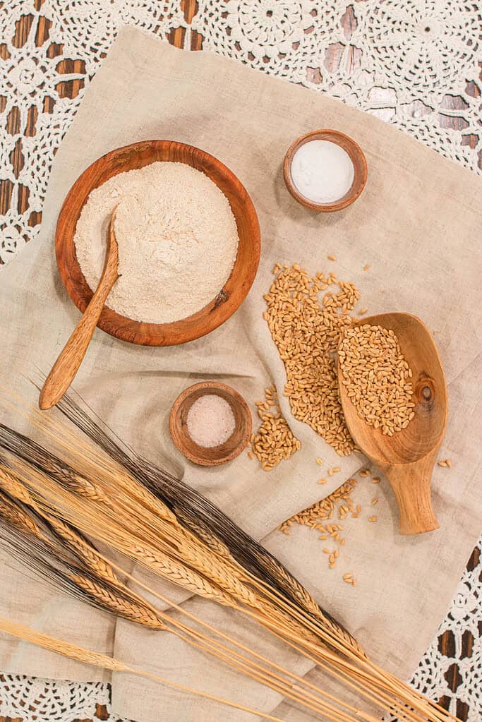 A rustic wooden bowl filled with freshly milled flour, surrounded by golden wheat kernels, stalks of wheat, a wooden scoop, and small bowls of salt and baking powder on a linen cloth.