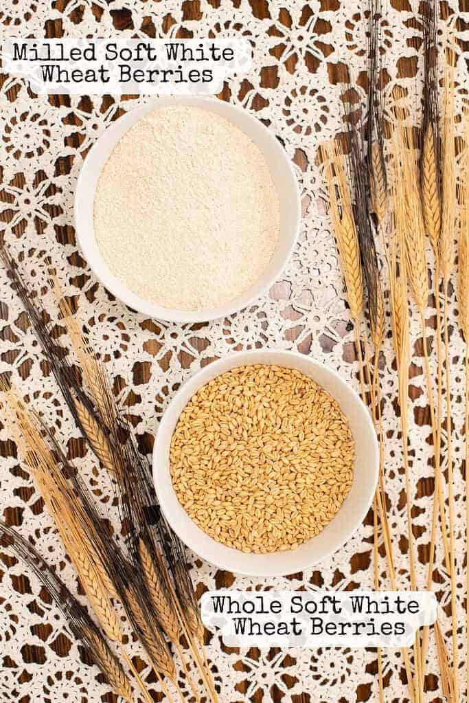 A white bowl of freshly milled soft white wheat flour is placed above a bowl of whole soft white wheat berries. The bowls are arranged on a delicate lace tablecloth with decorative stalks of wheat lying to the side.