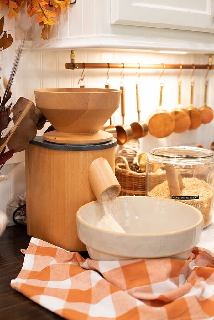 A wooden grain mill grinding hard white wheat into fresh flour, the flour falling into a cream-colored mixing bowl with a warm orange gingham cloth nearby.
