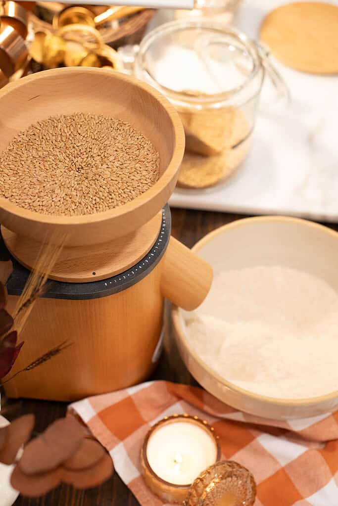 The hopper of a wooden grain mill filled with golden wheat berries, with a bowl of freshly milled flour placed beside it on a checkered cloth.