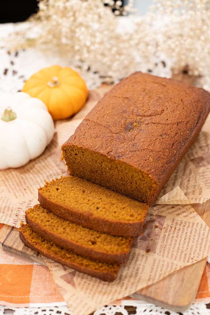 A golden-brown loaf of homemade pumpkin bread, sliced to reveal its soft, moist crumb, styled with mini pumpkins and lace in a cozy autumn setting.