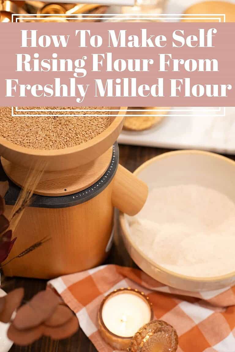 A cozy kitchen scene with a wooden grain mill filled with wheat berries, next to a bowl of freshly milled flour on a rustic table. An orange and white gingham cloth, a glowing candle, and dried florals add a warm cottage-style touch. Text overlay reads: “How To Make Self Rising Flour From Freshly Milled Flour.”