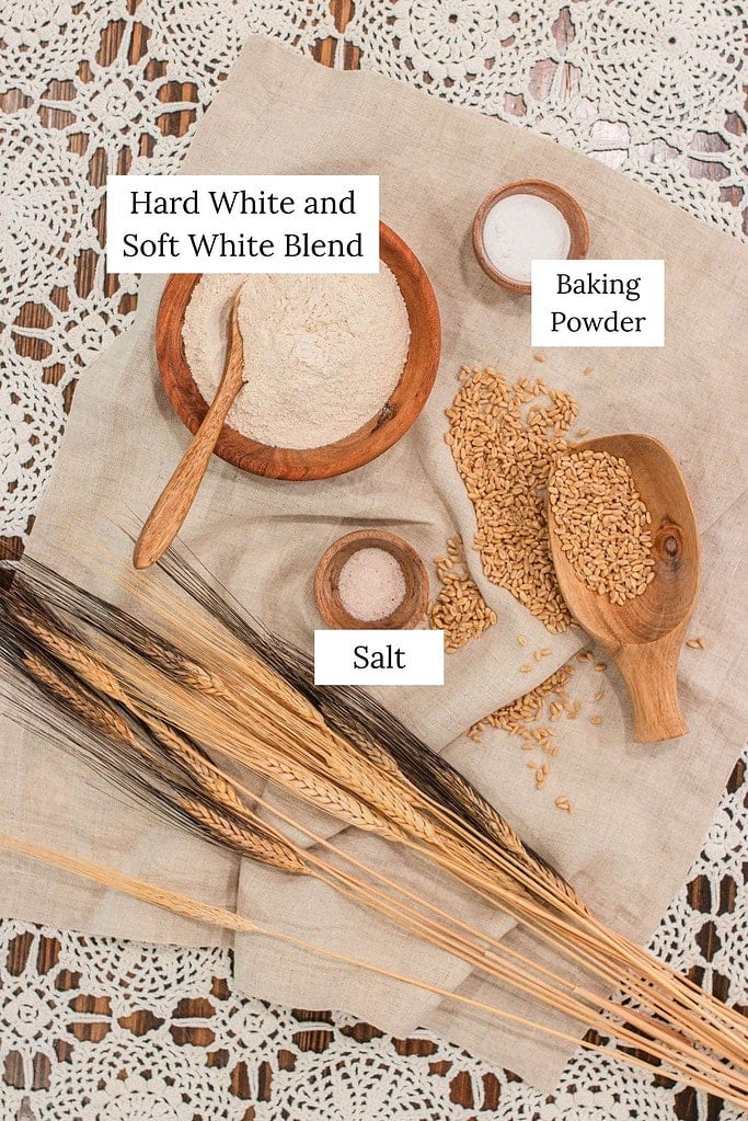 Top-down view of freshly milled flour in a wooden bowl labeled hard white and soft white blend, with small bowls of baking powder and salt, whole wheat berries in a wooden scoop, and wheat stalks arranged on a linen cloth over a lace tablecloth