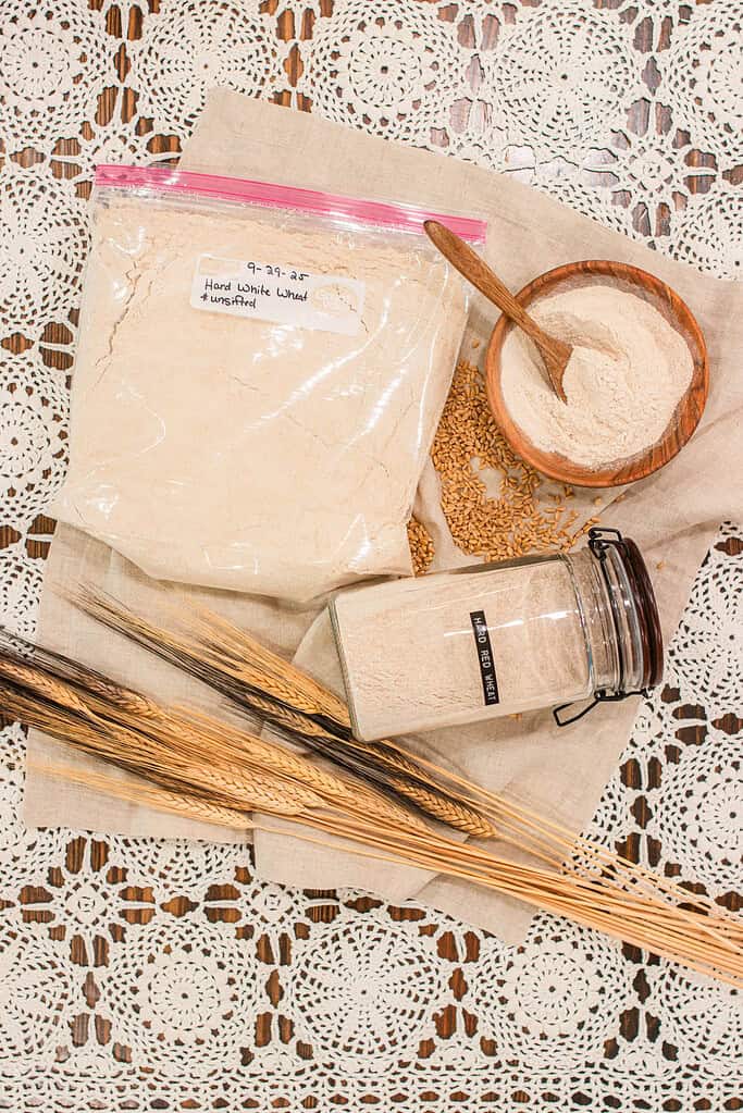 A cozy flat lay of freshly milled flour labeled Hard White Wheat, unsifted, stored in a clear zip bag beside a wooden bowl of flour, glass jar labeled Hard Red Wheat, scattered wheat berries, and stalks of golden grain on a linen cloth over a vintage lace tablecloth — illustrating how to store freshly milled flour.