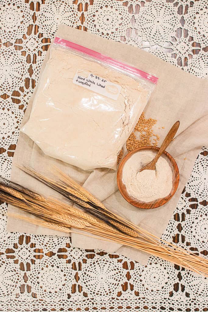 Freshly milled hard white wheat flour stored in a labeled plastic bag beside a small wooden bowl filled with flour and a spoon, loose wheat berries, and wheat stalks on neutral linen with a crochet lace background — showing proper short-term flour storage for home millers.